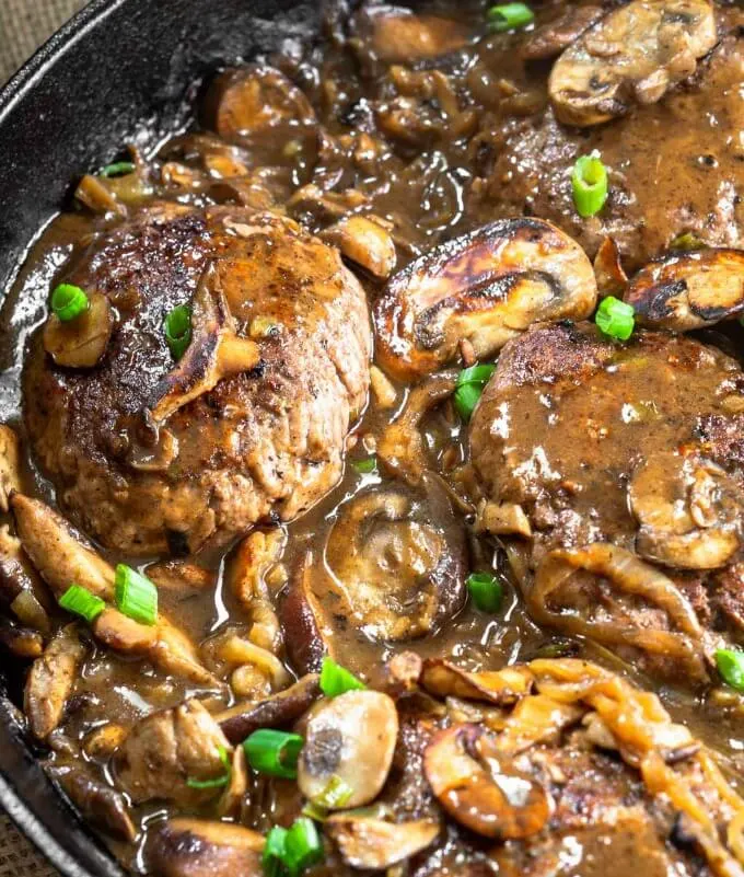 A Cast Iron Skillet full of Hamburger Steaks slathered in mushroom onion gravy and garnished with green onion