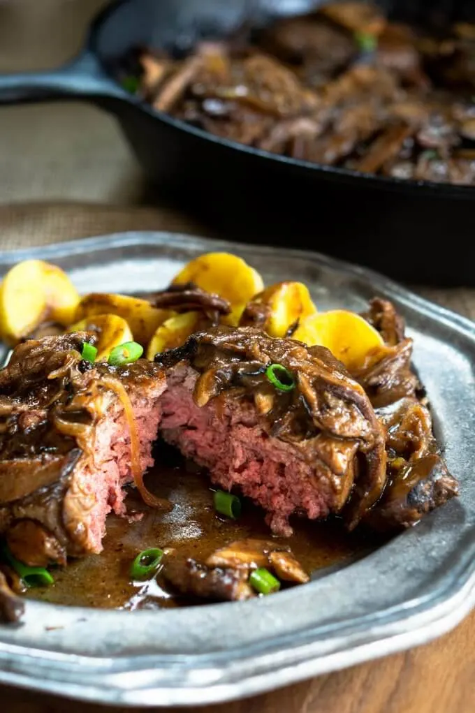 A cut open hamburger patty cooked to a pink medium rare smothered in brown gravy on a grey plate next to yellow gold potatoes