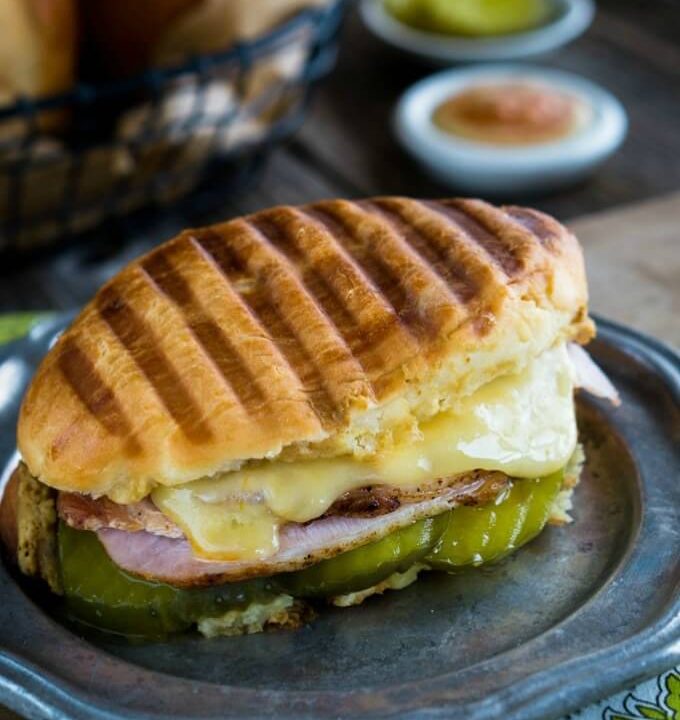 Oval Medianoche bread roll filled with dill pickle slices, ham, roasted pork, and swiss cheese on a grey metal plate. A white dish of a sriracha aioli sits in the background next to a basket of rolls.