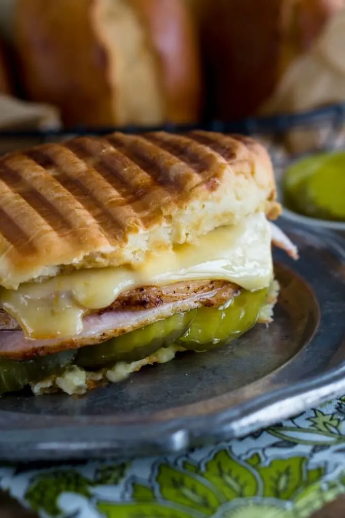 Close up of half a Cuban Sandwich made with grilled bread roll filled with pickles, ham, roasted pork, and Swiss cheese on a metal plate.