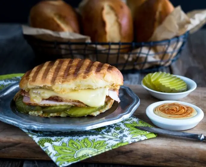 Oval Medianoche bread roll filled with dill pickle slices, ham, roasted pork, and swiss cheese on a grey metal plate and then grilled. White dishes of a sriracha aioli and pickle slices sit next to a basket of Medianoche rolls.