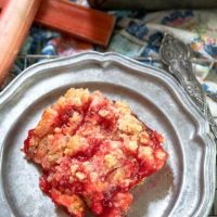 Rhubarb bar sitting on a metal plate in from of a pan filled with rhubarb dessert on top of a multi-color napkin and fresh rhubarb in the background.