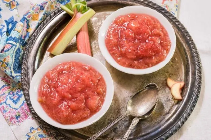 Top view of two white bowls filled with rhubarb sauce on an antique tray with old spoons and fresh rhubarb stalks