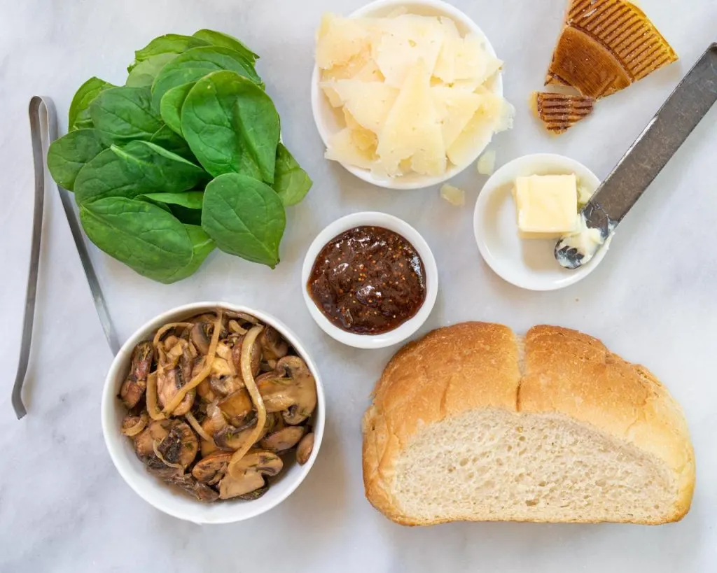 Top down view of ingredients used to maake a Manchego grilled cheese sandwich including bread, cheese, spinach, onion, mushrooms, and fig spread.