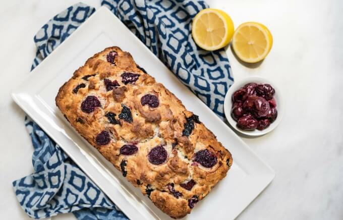 A top-down loaf of cherry bread sits on a white plate over a blue patterned plate. A small white bowl of cherries and to half lemons sit next to the plate.