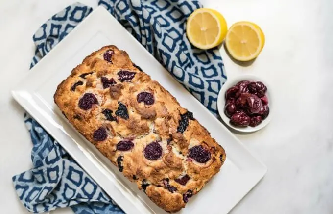 A top-down loaf of cherry bread sits on a white plate over a blue patterned plate. A small white bowl of cherries and to half lemons sit next to the plate.