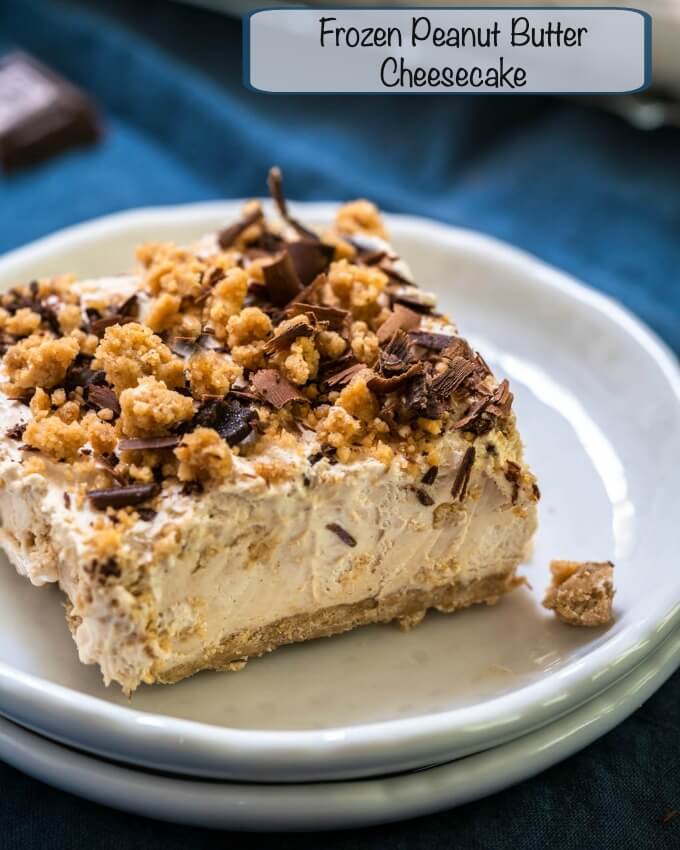 A square piece of peanut butter cheesecake topped with crumble and shaved chocolate sitting on a stack of two white plates and a blue napkin.