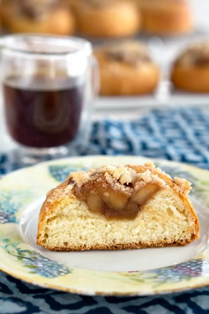 Pastry roll filled with apple filling cut in half sitting on a floral plate. A cup of coffee and more rolls sit behind the plate.