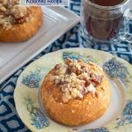 A browned kolache roll filled with baked apples and topped with a cinnamon crumble sitting on a floral plate and blue napkin. A cup of coffee and a tray of rolls sit behind it.