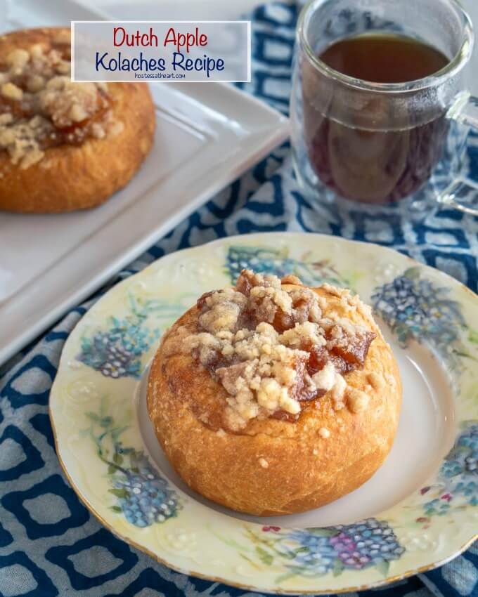A browned kolache roll filled with baked apples and topped with a cinnamon crumble sitting on a floral plate and blue napkin. A cup of coffee and a tray of rolls sit behind it.