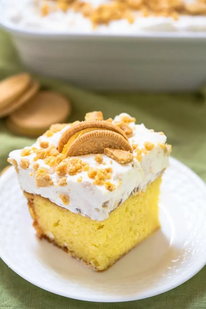 Slice of yellow lemon cake topped with white whipped topping, crushed cookies and a half cookie on a white plate. A white baking dish and whole cookies sits behind it.