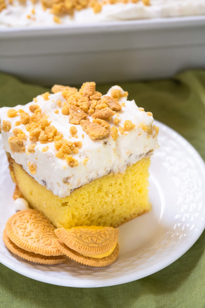 Bright yellow slice of cake with whipped topping and crushed cookies on a white plate with two broken cookies sitting on the same plate.