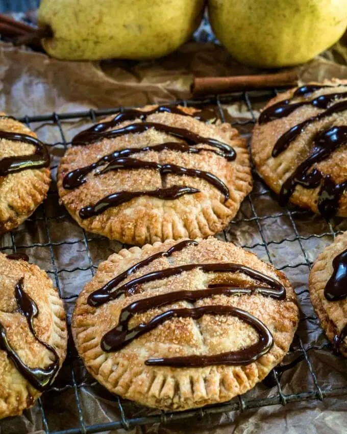 Two round small round pies baked to a golden brown with dark chocolate drizzled over the top sitting in front of two fresh green pears