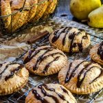 A baking rack topped with 6 pear hand pies baked brown and drizzled with chocolate. A basket of more pies and raw pears sit in the background.