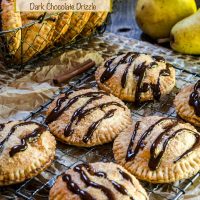 A baking rack topped with 6 pear hand pies baked brown and drizzled with chocolate. A basket of more pies and raw pears sit in the background.