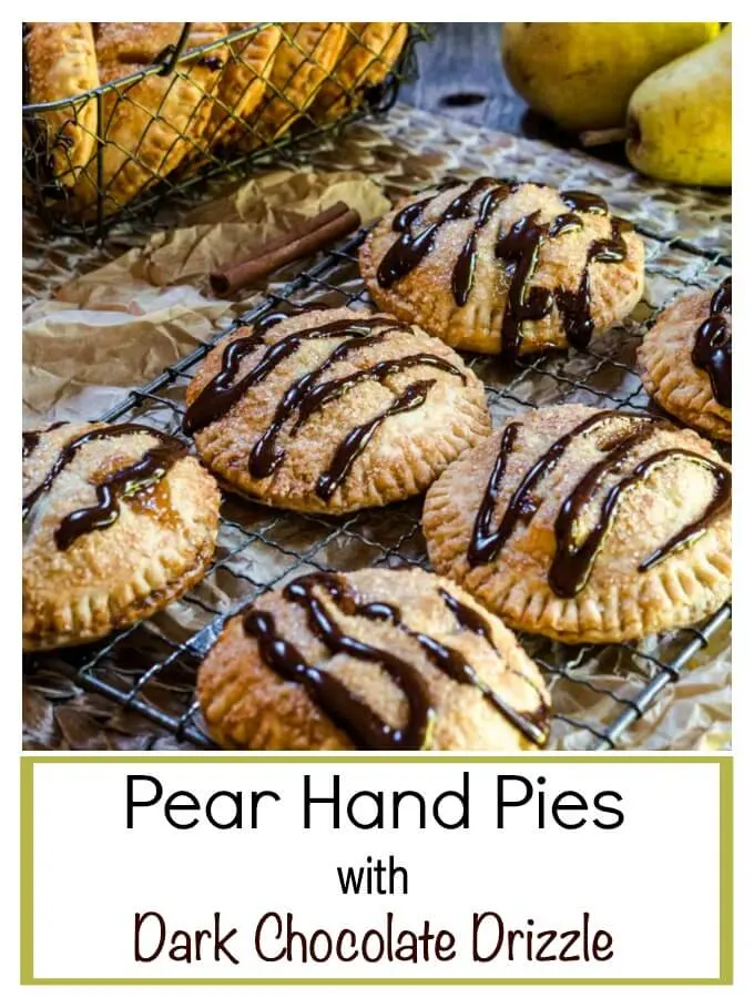 A baking rack topped with 6 pear small 4 inch pies baked brown and drizzled with chocolate. A basket of more pies and raw pears sit in the background.