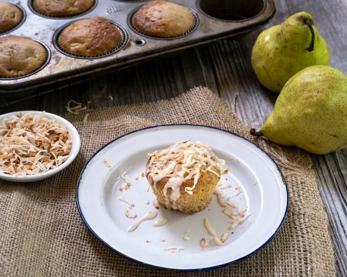 Single muffin topped with coconut on a white plate surrounded by fresh pears, coconut in a bowl and a tin of muffins