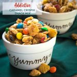Close up of Crispix snack mix in a white ramekin with "yummy" printed on the front. The mix is full of Crispix cereal, M&M's and peanuts sitting on a green napkin dotted with spilled snack mix. another ramekin full of snack mix sits in the background. The title "Addictive Caramel Crispix Snack Mix" is printed in the top left of the photo.