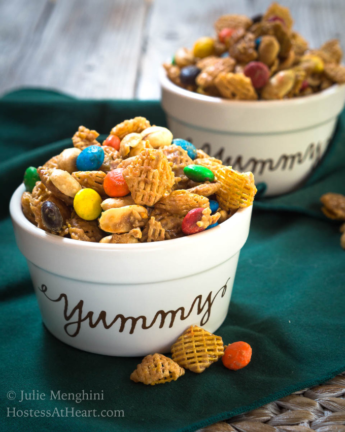 Zwei Schüsseln mit dem Wort \"yummy\" auf der Vorderseite sind überfüllt mit Crispix-Müsli, Erdnüssen und MM-Mix, die auf einer grünen Serviette mit verschütteten Müslistücken sitzen."yummy\" on the front are overflowing with Crispix cereal, peanuts and M&M mix sitting on a green napkin with pieces of cereal spilled in front.