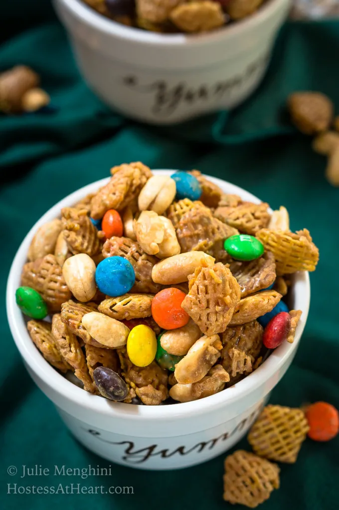Top down view of an overflowing bowl of brown sugar glazed Crispix cereal, peanuts and m&ms sitting on a green napkin and spilled cereal mix.