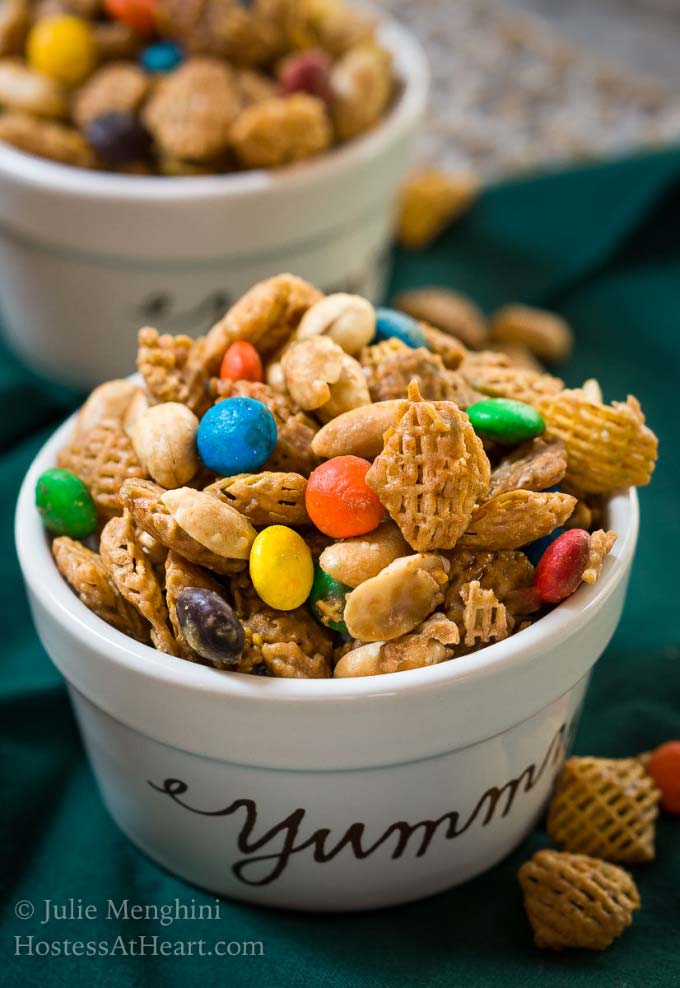 Close up of Crispix snack mix in a white ramekin with \"yummy\" printed on the front. The mix is full of Crispix cereal, M&M\'s and peanuts sitting on a green napkin dotted with spilled snack mix. another ramekin full of snack mix sits in the background.
