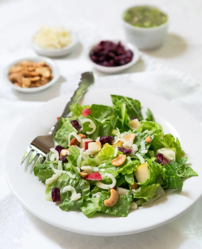 Angled photo of a green salad topped with apples, pears, cheese, cashews and craisins. Small white bowls hold extra dressing, cashews, cheese and craisins behind the salad bowl.