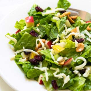 Close up of a lettuce salad in a white bowl topped with cashews, cranberries, pears, apples and grated swiss cheese