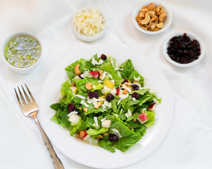 Top-down photo of a lettuce salad in a white bowl topped with cashews, cranberries, pears, apples, and grated Swiss cheese. Small bowls of dressing, cheese, cashews, and cranberries sit in the background.