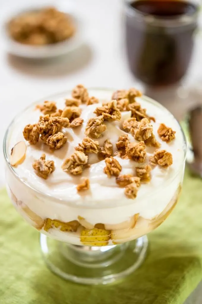 Top view of a glass dish of yogurt layered with pears and sprinkled with granola on a green napkin. Coffee cup sits behind it.