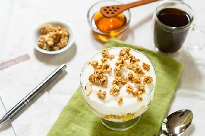 A table set with yogurt topped with granola on a green napkin next to a coffee cup, spoon, dish of granola and a shopping list topped with a pen.