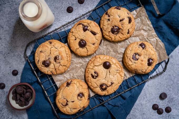 Top-down shot of 6 chocolate chip cookies cooling on a rack over a blue towel next to a jar of milk and a small bowl of chocolate chips.