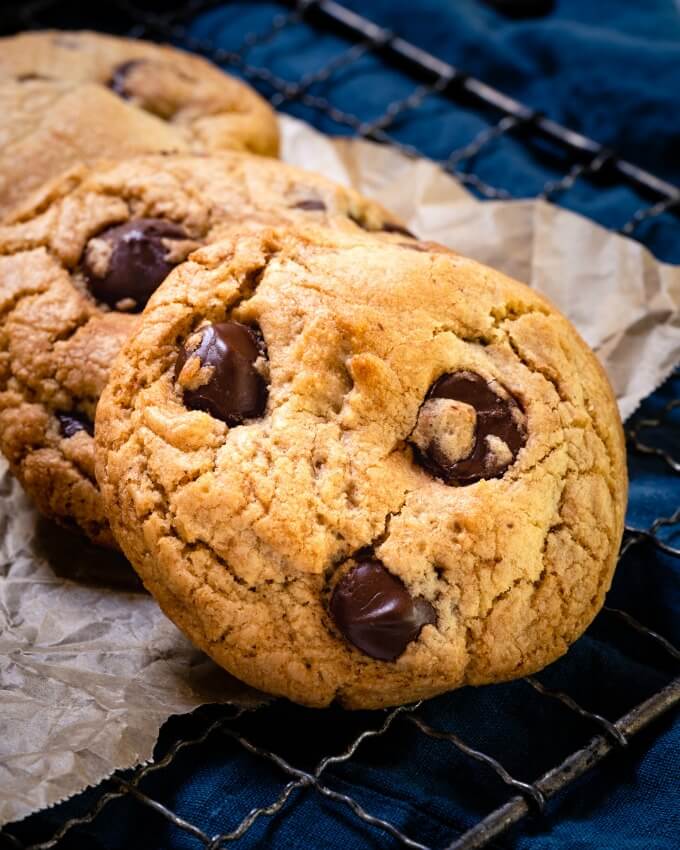 Close up of 3 layered chocolate chip cookies dotted with large pieces of dark chocolate sitting on a rack over a bue towel.