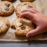 6 chocolate chip cookies on parchment paper with a childs hand returning one with a bite taken out of it.