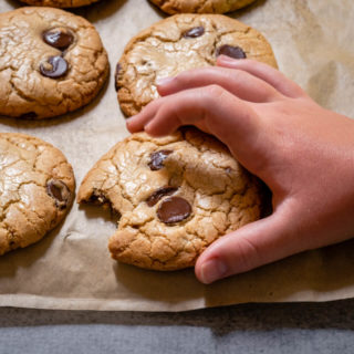 6 chocolate chip cookies on parchment paper with a childs hand returning one with a bite taken out of it.