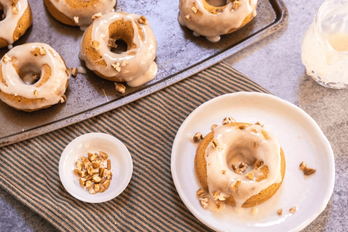 Top shot of a pumpkin donut on a white plate sitting on a striped towel with a pan of donuts, bowl of pecans sitting next to it.