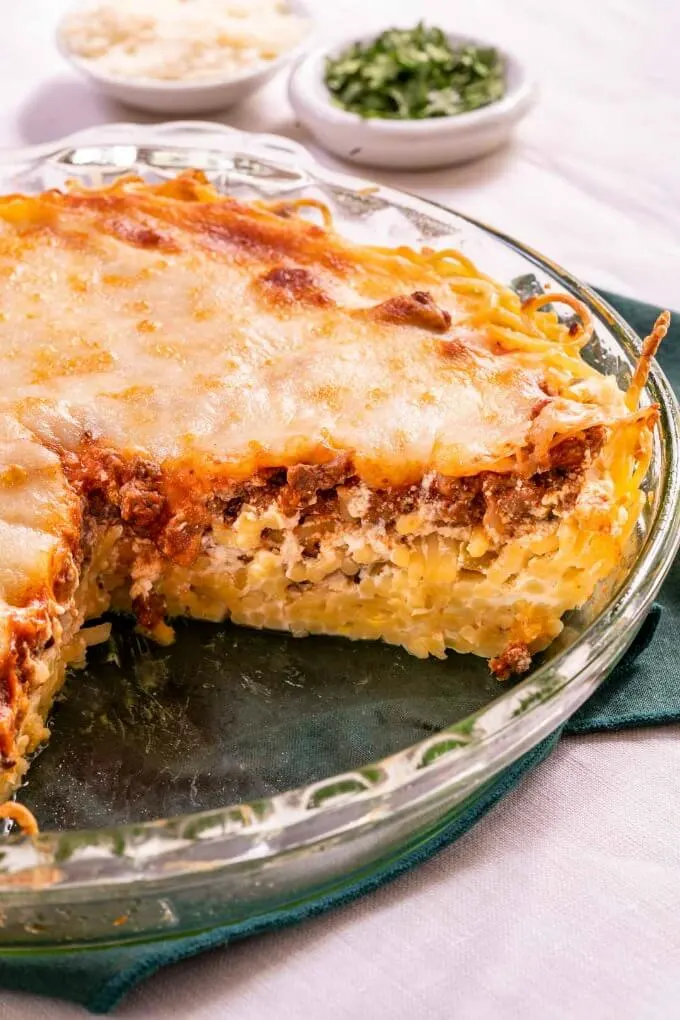 A pie plate with baked spaghetti pie with a cut piece removed showing layers of pasta, meat sauce and mozzarella.
