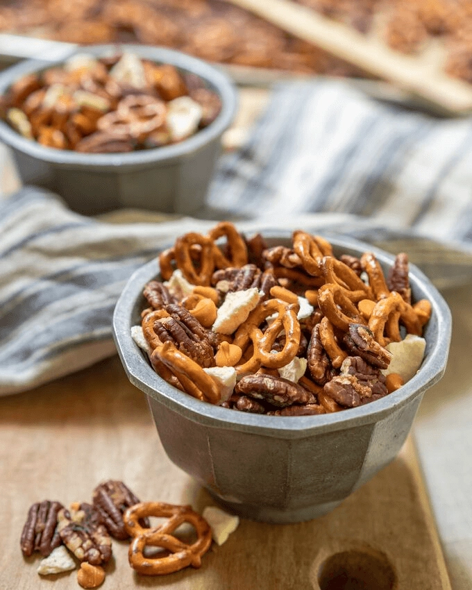 Top angle view of two bowls of a snack mix of pretzels, pecans, caramel chips, and dried apple sitting on a wooden board and a blue towel. Spilled mix lies next to the bowl.