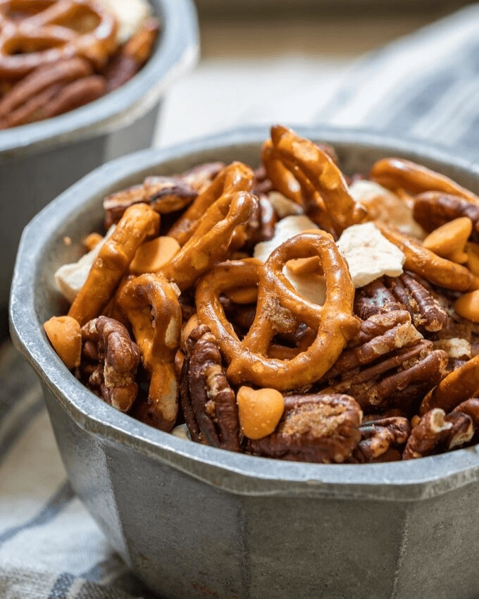 Side view of a snack mix of pretzels, pecans, caramel chips and dried apples in a metal bowl.