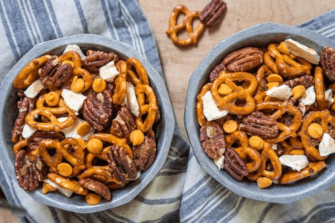 Top view of two bowls filled with a snack mix of pretzels, pecans, caramel chips, and dried apples on a blue striped napkin