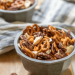 Top angle view of two bowls of a snack mix of pretzels, pecans, caramel chips, and dried apple sitting on a wooden board and a blue towel. Spilled mix lies next to the bowl.