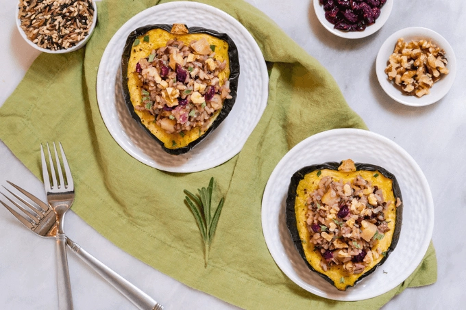 Top photo of two haves of Acorn Squash stuffed with Wild Rice and apple. Small bowls of rice, nuts and crasins sit in the back ground.
