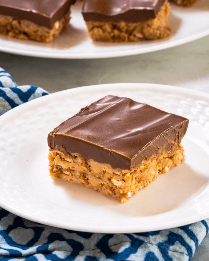 Peanut Butter bar topped with thick creamy chocolate layer sitting on a white plate in front of a serving platter with more dessert squares on it.