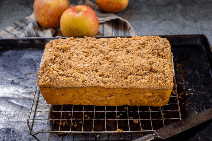 Loaf of pumpkin bread sitting on a rack over a baking sheet with 3 apples sitting behind it on a striped towel.