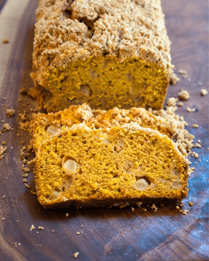 Slice loaf of pumpkin bread dotted with chunks of apple and topped with streusel topping sitting on a wooden cutting board