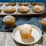 Angle photo of a powder sugar dusted pumpkin muffin sitting on a white plate surrounded by other muffins in front of a tin of muffins.