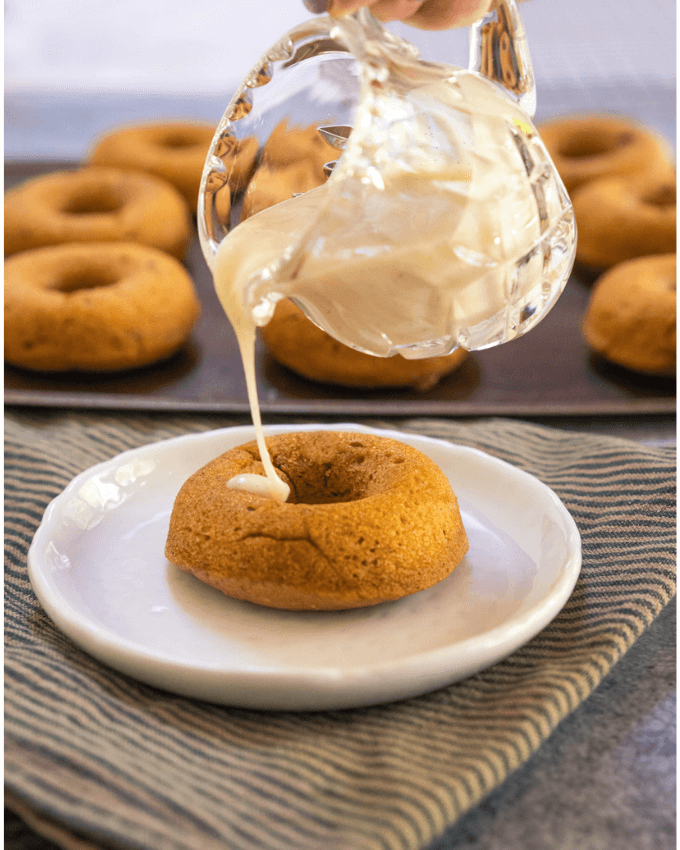 Glass pitcher pouring glaze on a pumpkin donut sitting on a white plate. A pan of donuts sit behind it.