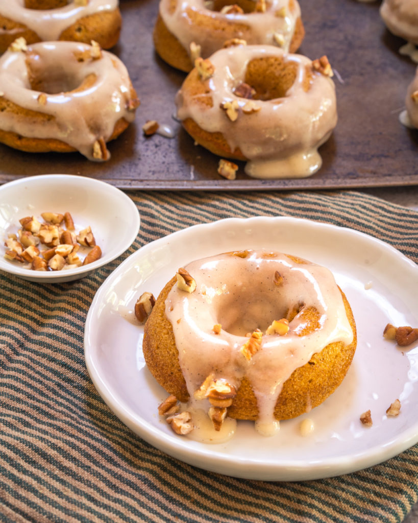 Donut with glaze and pecan pieces sitting on a white plate on a stripped towel. A pan of donuts and a dish of pecans sit behind it.