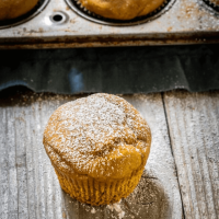 Single pumpkin muffin dusted with powdered sugar sitting on a wooden board in front of a tin of muffins