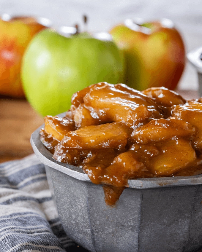 3/4 view of cooked apple pie filling in a metal bowl with 3 fresh apples behind it sitting on a blue stripped towel.