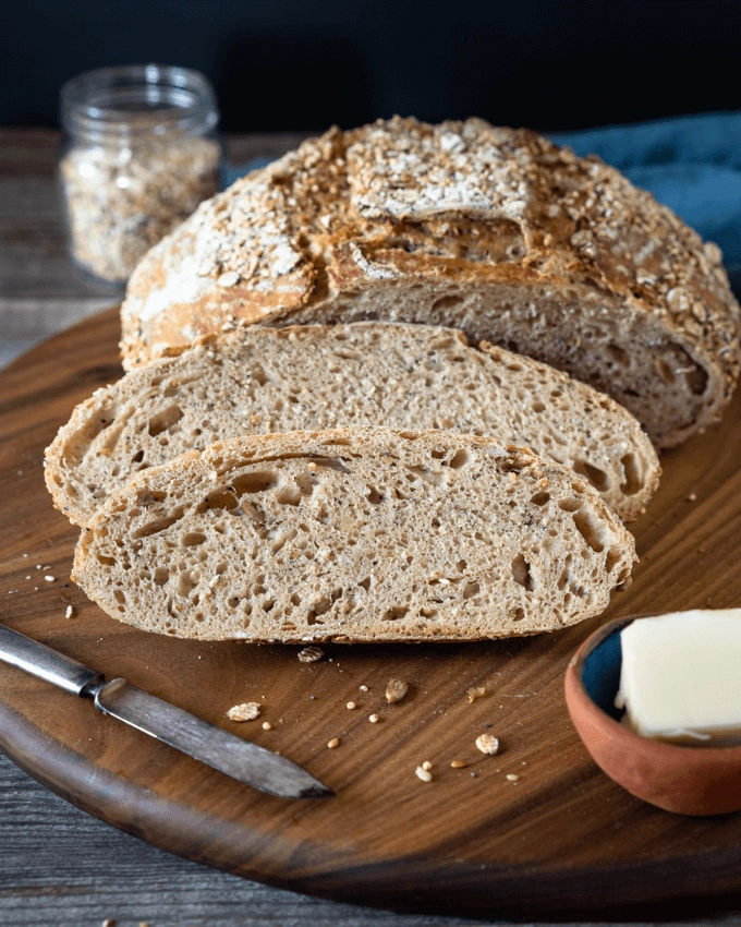 A round loaf of bread and two slices sitting up against it on a wood cutting board with a small cup holding butter and a small knife. A jar of grain and seeds sit behind it on a blue towel.